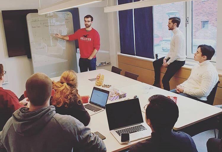 A meeting room. Mattias Bergström, a young man i a red shirt is holding a presentation pointing at a whiteboard. Five people are sitting around a table listening. One guy is leanign against the window sill.