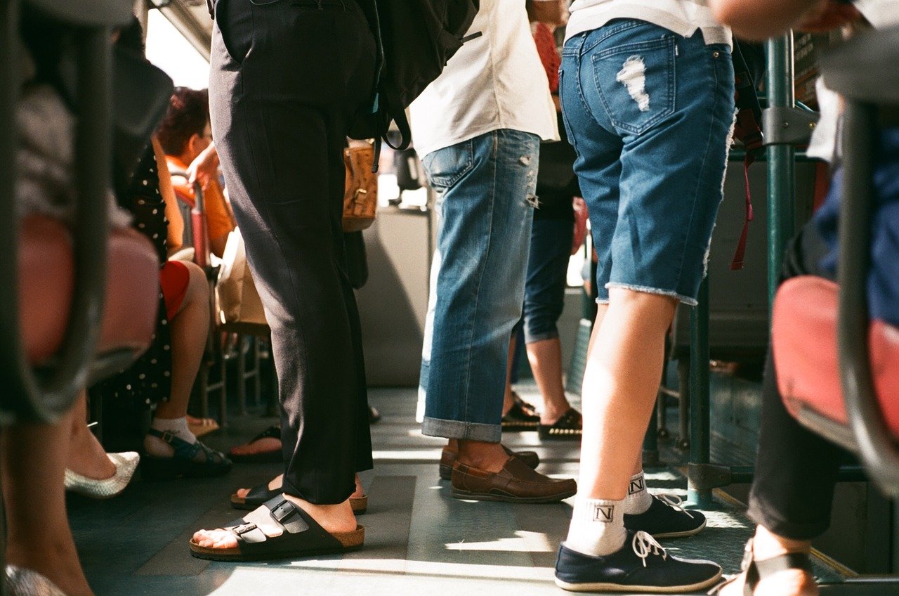 A full train cart, where you see the legs and feet of the passengers. Three people are in focus; one with black pants and birkenstock, one with jean shorts and sneakers and one with jeans and loafers. The sun shines in from the left.