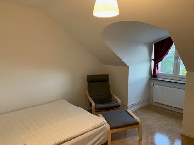 Bed with mattress cover, grey armchair, window with pink curtains in white painted apartment.