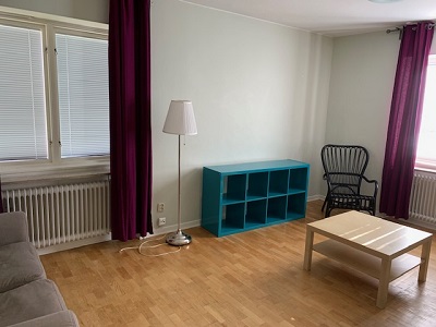 White painted living room with purple curtains, lamp, sofa, chair, bookcase and table.
