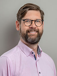 En man med kort mörkt hår, skägg, glasögon och rosa skjorta.