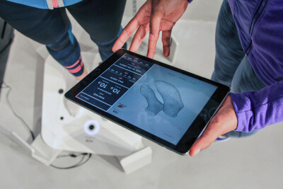 En hand som håller en iPad där ett par 3D scannade fötter visas. I bakgrunden fötter på 3D scanner.