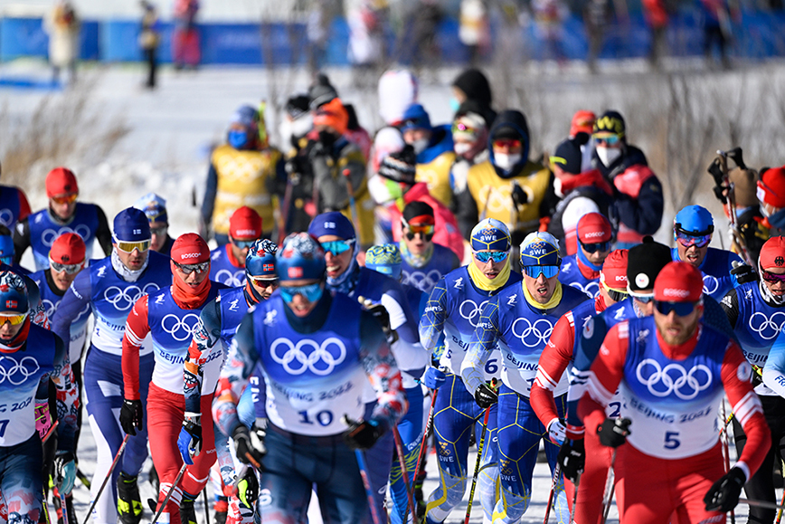 Ett 20-tal skidåkare i spåret med publik i bakgrunden under en tävling i olympiska spelen.
