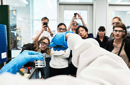Händer i blå platshandskar håller upp en bägare. Människor i labbmiljö tittar på. 