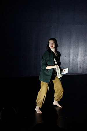 En dansare i beiga byxor, svart tröja som rör sig i ett mörkt rum.