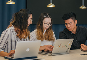 Three students talking at a table looking while at their computers