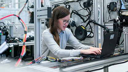 Kvinna som sitter vid en dator i en labbmiljö med sladdar och teknisk utrustning.