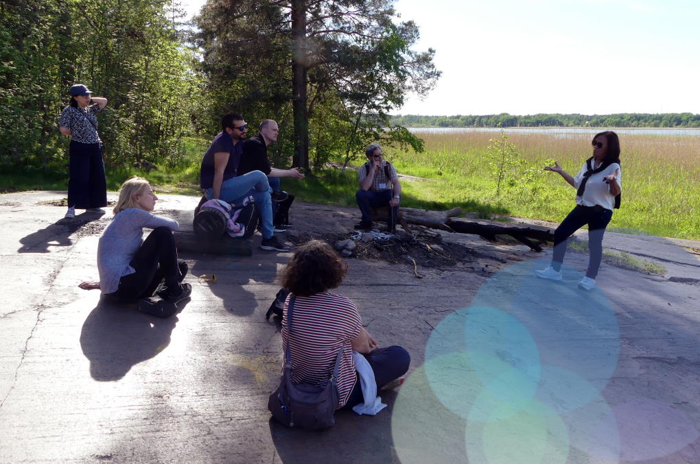 En grupp människor sitter utomhus på en klipphäll en sommardag och lyssnar på en föredragshållare.
