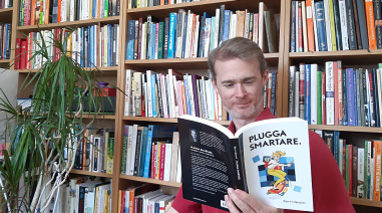 Lecturer Björn Liljeqvist reading a book while leaning against a bookshelf filled with books