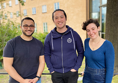 Mustafa Al-Janabi, Frank Jiang and Elisa Bin outside in front of shrubbery and a yellow building.