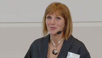 A blond woman with a large silver necklace and grey jacket.