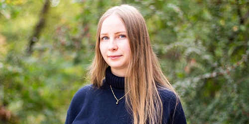 En kvinna med blont hår och mörkblå tröja står framför en grönskande bakgrund. Hon är KTH-student.