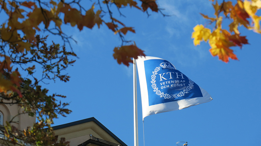 KTH flag with blue university seal on white field, unfurls in wind, with autumn leaves in foreground