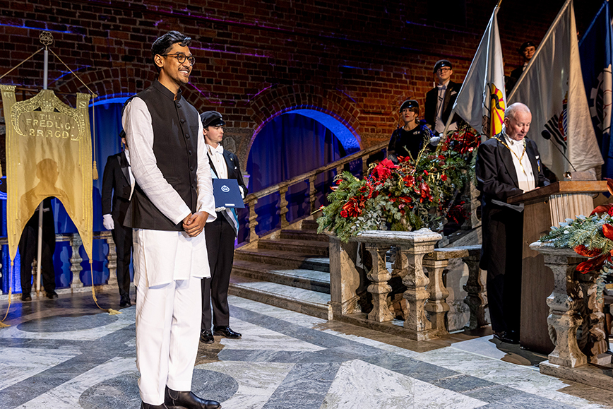 Man stands on stage at graduation ceremony, while president of KTH speaks at lectern. 