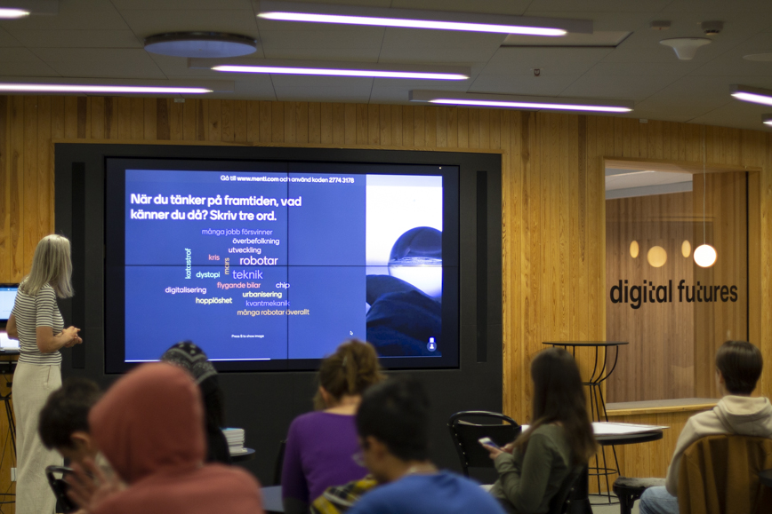 Elever i skolbänkar i förgrunden framför en stor skärm.