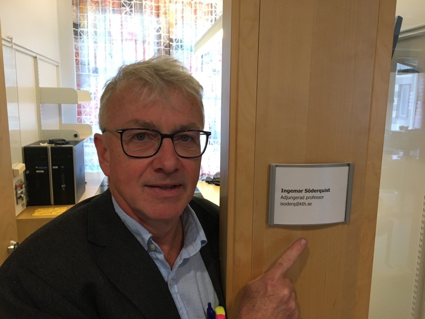 Man looking into the camera pointing at name tag on door. 