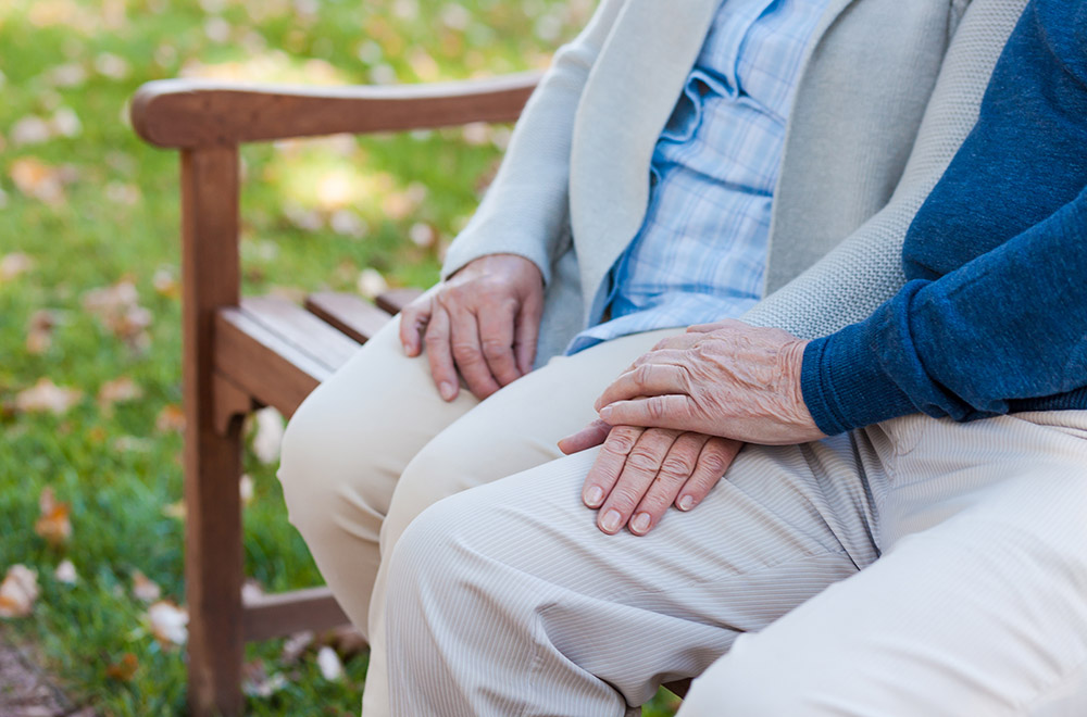 Ett äldre par sitter på en parkbänk, delar av överkropparna och benen samt händerna återfinns i bild