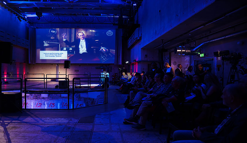 En person på scen framför en stor skärm och framför publik som sitter i mörker.