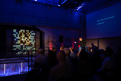 A dark venue with blue spotlights and an audience facing a stage where a person is.