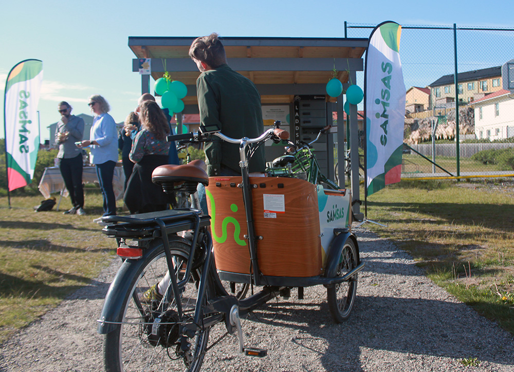 A box bike in front of a bicycle garage, next to it several people are listening to speakers.