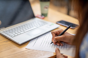 Person writing on print-out document. Laptop in background.