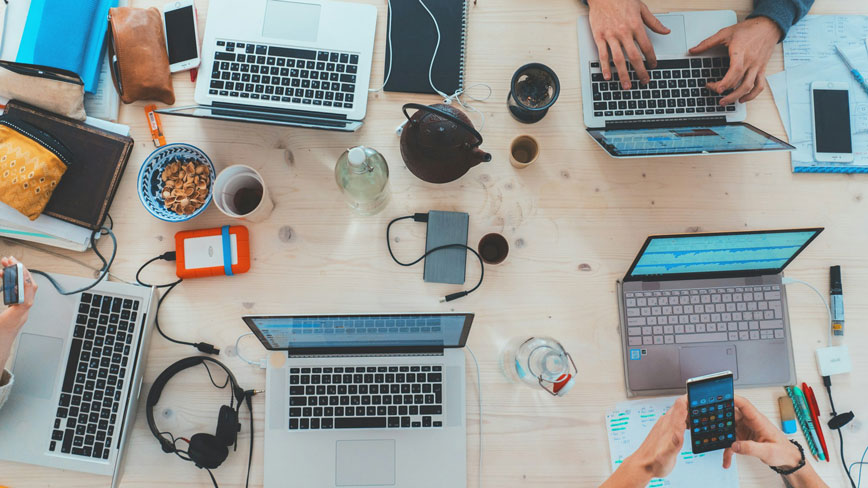 People sitting down near table with assorted laptop computers, phones, notepads and coffee.