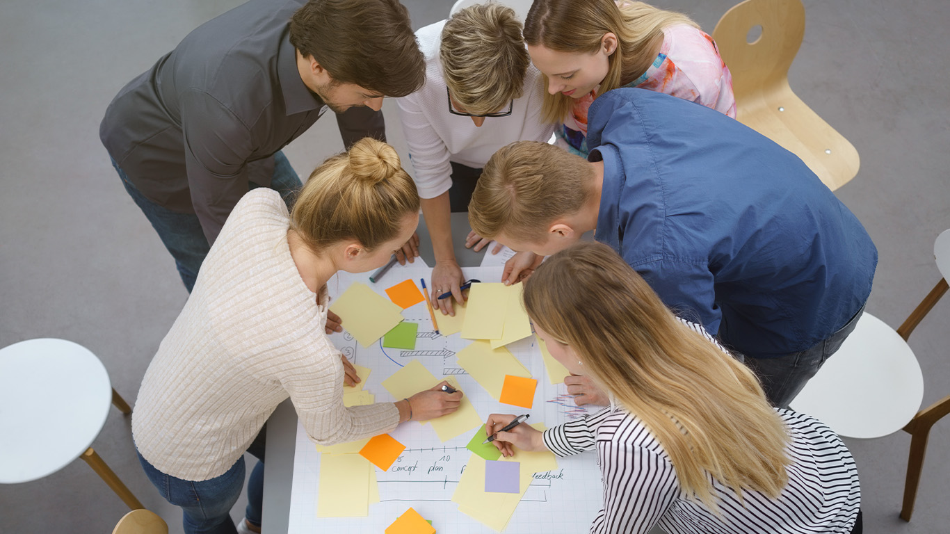 Personer som står nedåtböjda över ett bord med post-it och skisser.