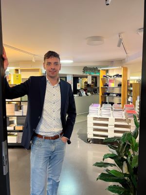 Person in doorway of store with shelves and paper stacks in background.