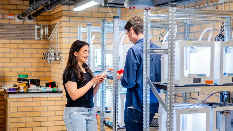 Two students standing beside shelves full of 3D printers