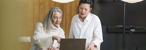 Två kvinnor i vit tröja respektive blus pekar och tittar ihop på en laptop, i en stor ljus lokal.
