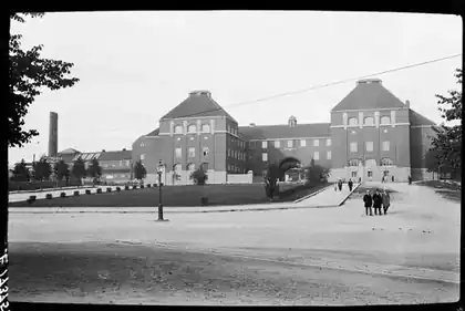 Svartvitt foto av KTH:s huvudbyggnad i tegel med två markanta gavelförsedda flyglar, sett över en öppen plats där några personer syns. Till vänster reser sig en skorsten.