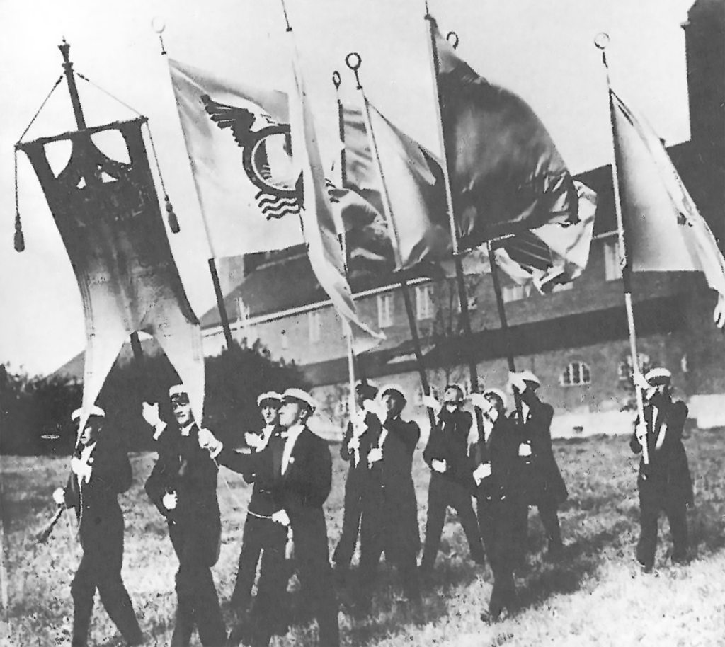 Students in dark suits and white student caps march across a lawn carrying several flags and a banner, with a brick building in the background.