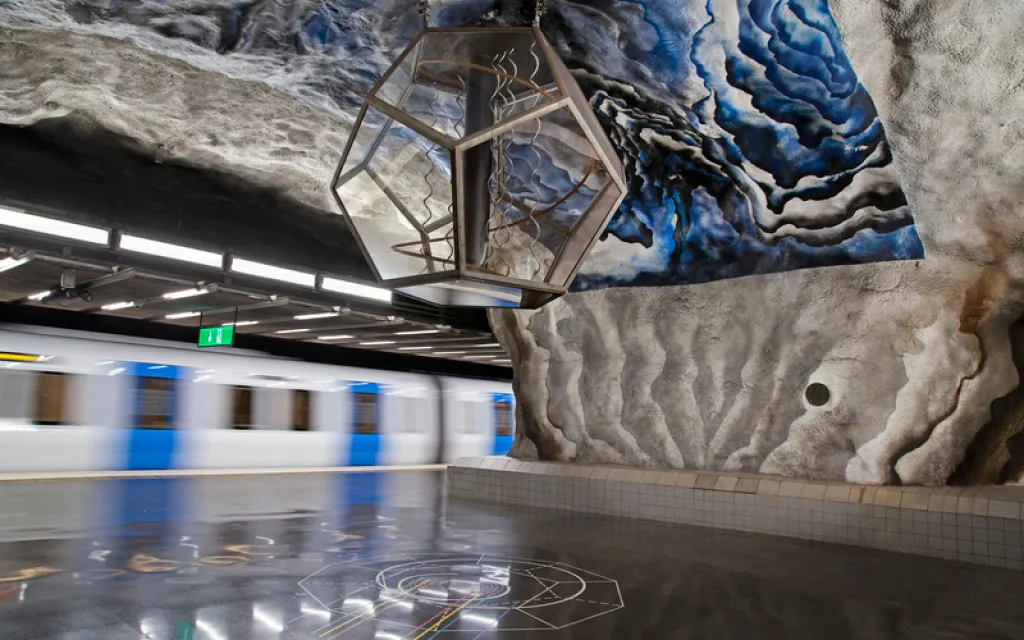 Platform of a metro station carved from rock. A large glass dodecahedron hangs from the ceiling, the walls are painted in blue and white, and a blue-and-white train passes on the left.