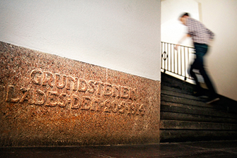 Photo: a wall with reddish foundation stone at the bottom. A person walks up stairs connected to it.