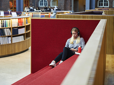A girl sits in the library, listening to a book, wearing headphones.