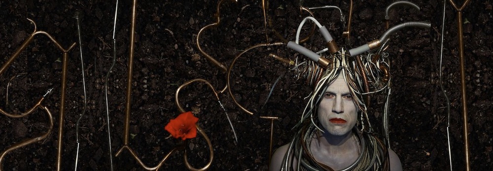 En screenshot från urkarnarsvarld.se: En man med vitmålat man bär en typ av huvudbonad gjord av olika typer av urkopplade rör och kablar.