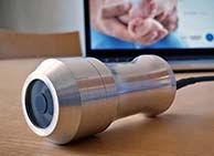 The jaundice meter is silver, round and about five centimeters long. A cord comes out of the back. In the background of the image is a computer displaying a picture of a baby.