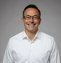 Donnie Lygonis, en medelålders man, ler mot kameran iklädd vit skjorta. Bilden är tagen mot en grå bakgrund. 